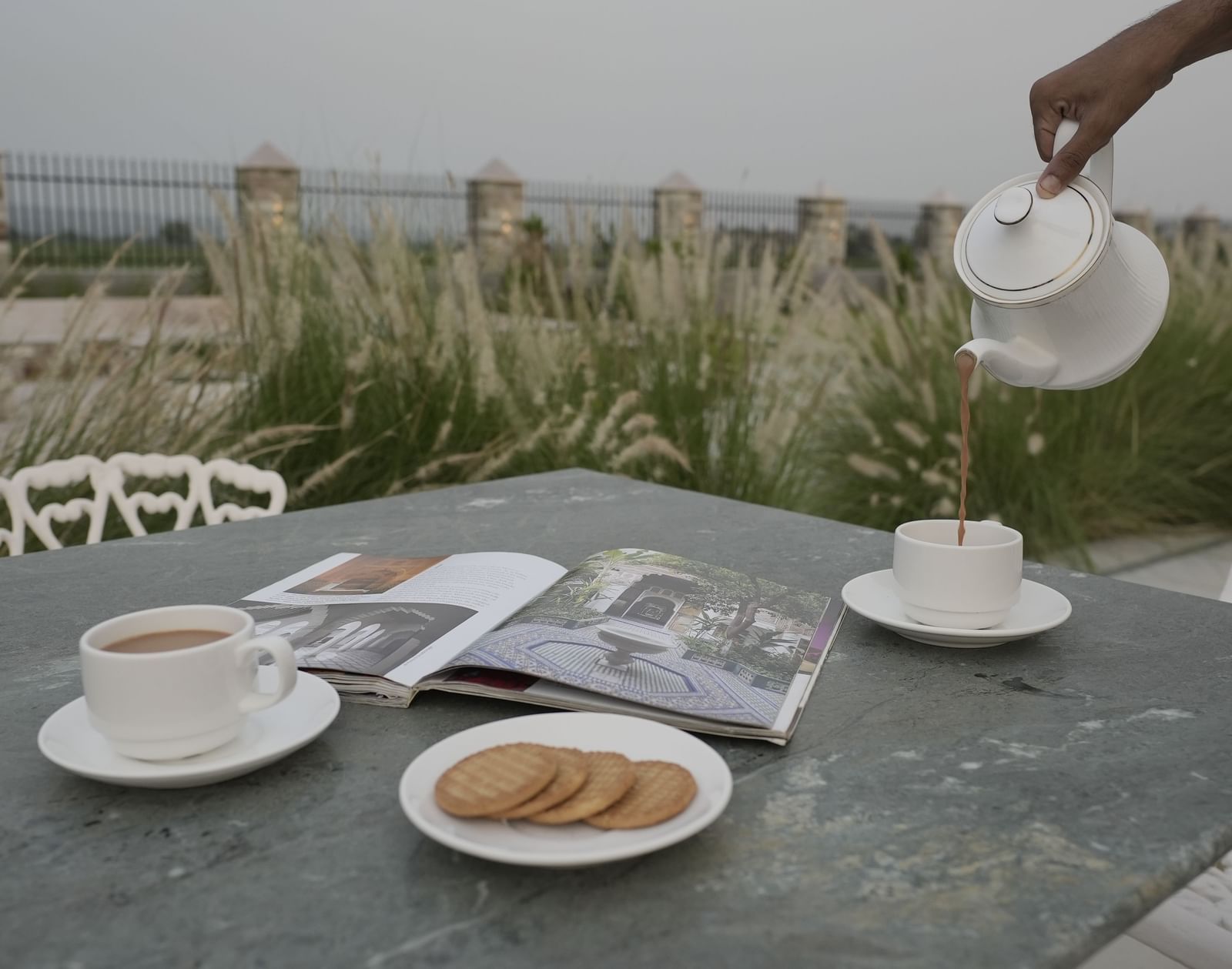 A close-up shot of an outdoor table at Mahendra Niwas with two cups of tea, a plate of biscuits, and a magazine. A person's hand pours tea from a white teapot into one of the cups.