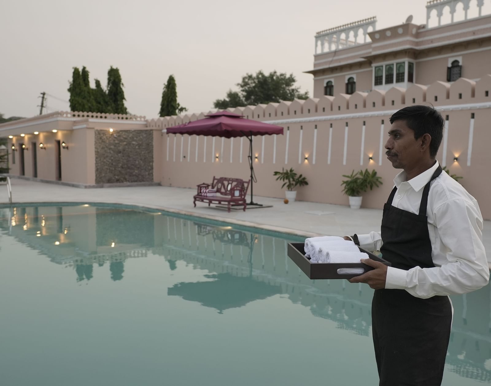 A close-up shot of a server in a white shirt and black apron holding a tray of white towels next to a large pool at Mahendra Niwas.