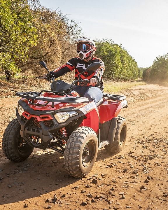 A man quad biking featuring full biking gear, dusty road, and surrounded by tall green trees.