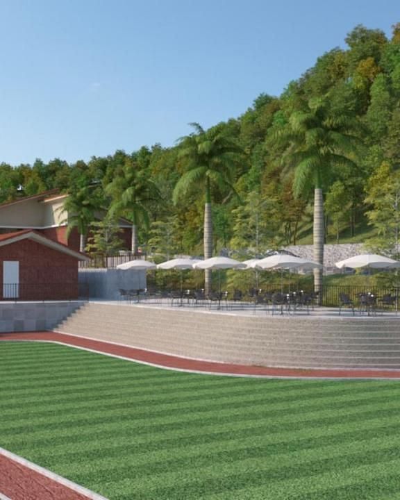 A red-roofed building with a green lawn in front, featuring a seating area with white umbrellas, tables, and chairs, set against a backdrop of trees and a hillside - Gaurav Lords Resort Shrivardhan. 