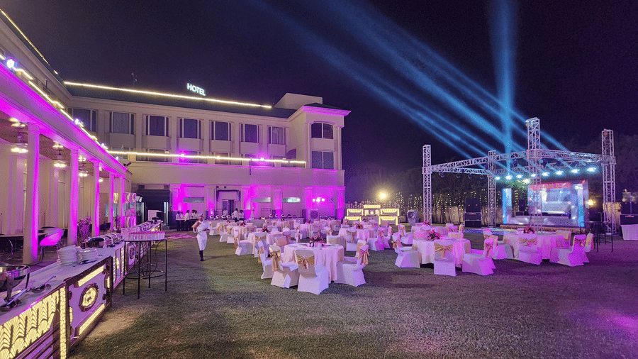 Wide view of the lawn at Ambrosia Sarovar Portico, Haridwar, prepared for a night celebration with round banquet tables, buffet setup, stage lighting, and a lit facade.