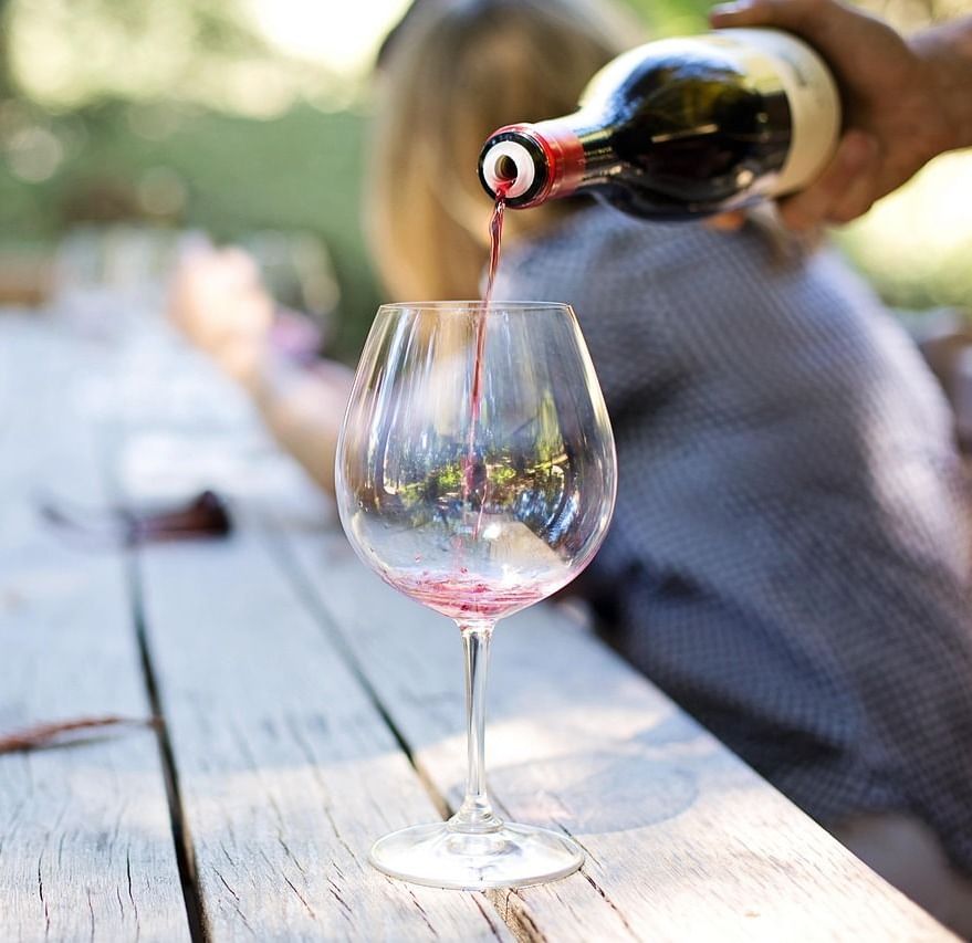 A hand pouring red wine from a bottle into a clear wine glass placed on a rustic wooden table outdoors, with blurred people in the background.