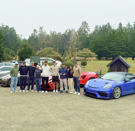 Group of people standing near several parked cars in an open area surrounded by tree.