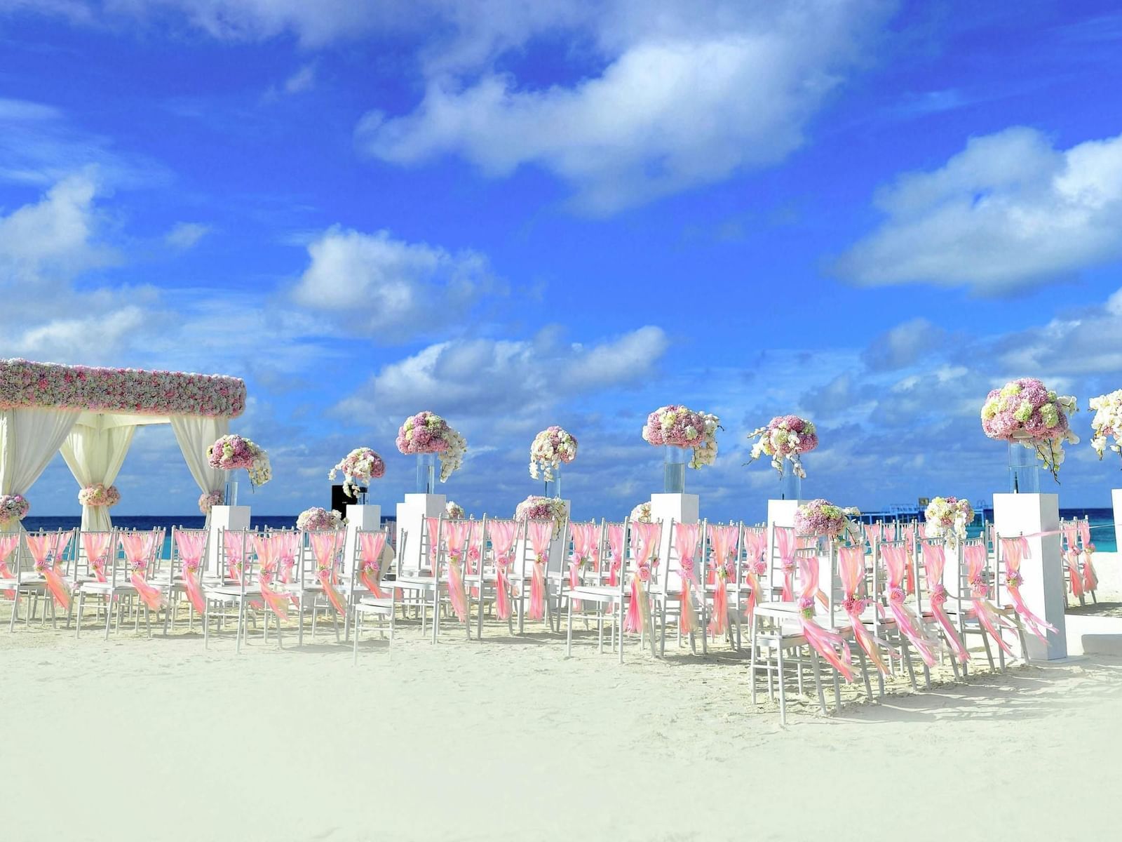 Side view of a wedding setup with neatly arranged pink and white chairs facing an altar by the beach under a clear blue sky.