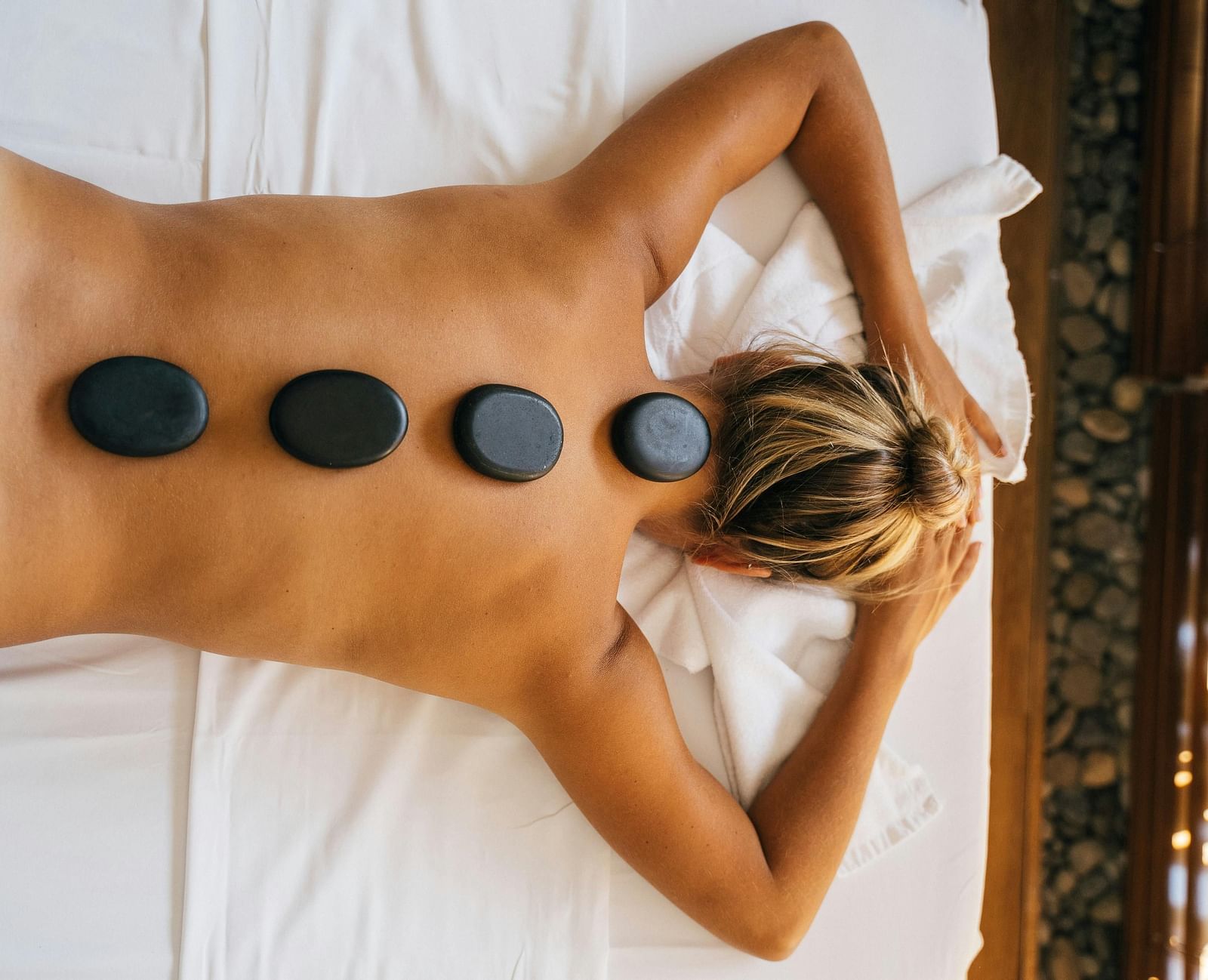 A woman lying down with back with black stones kept over her spine.