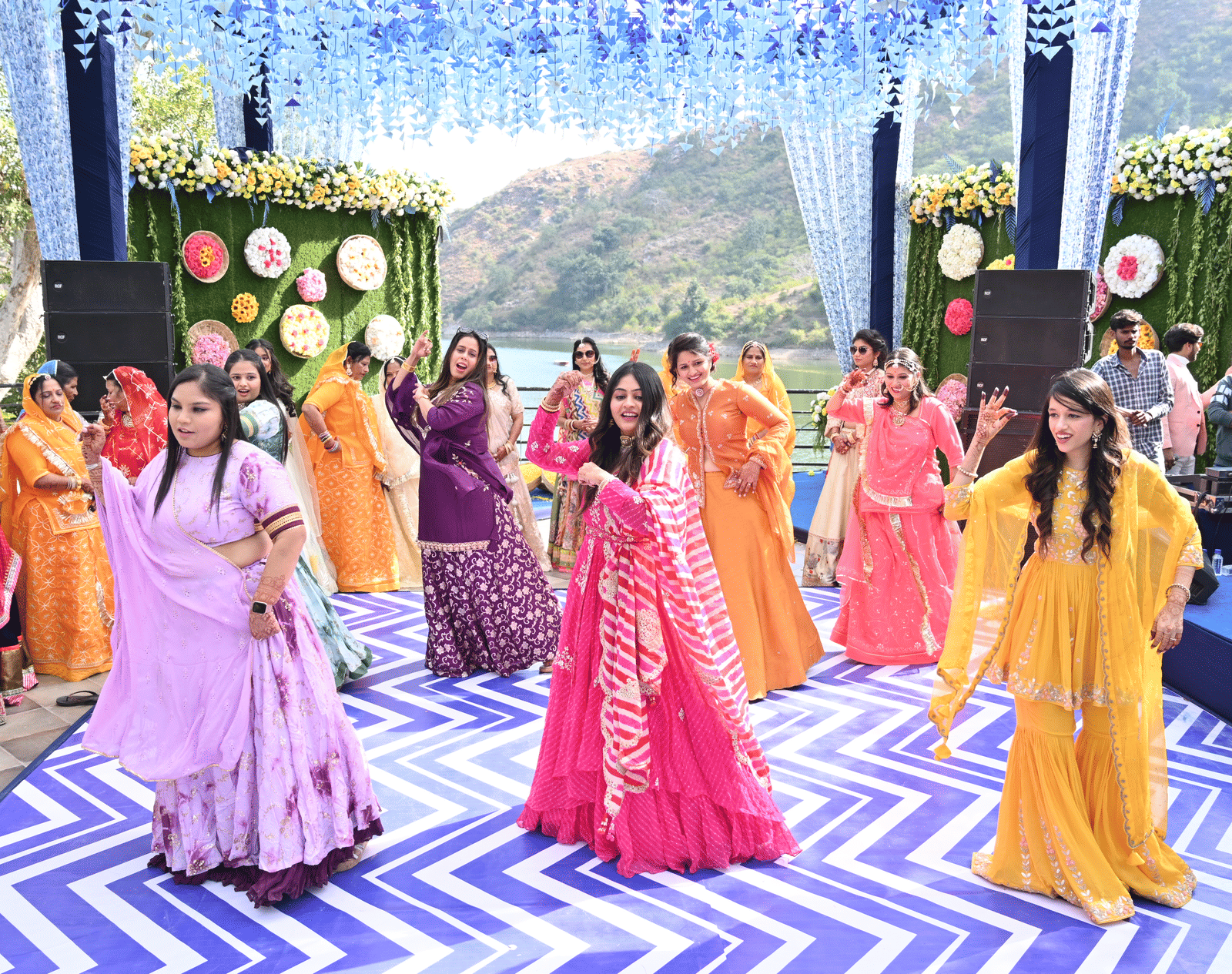 Women in colourful traditional outfits dancing at a daytime outdoor celebration, with floral decor at a Lakeview wedding venue, Kumbhalgarh - Via Lakhela Resort & Spa.