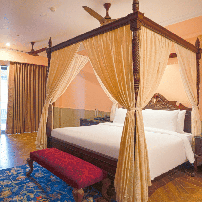 A bedroom with a four-poster bed draped in sheer curtains, wooden flooring, cushioned bench, sliding curtains and a small dining table set near the window at MAYFAIR Bay Resort, Paradeep.