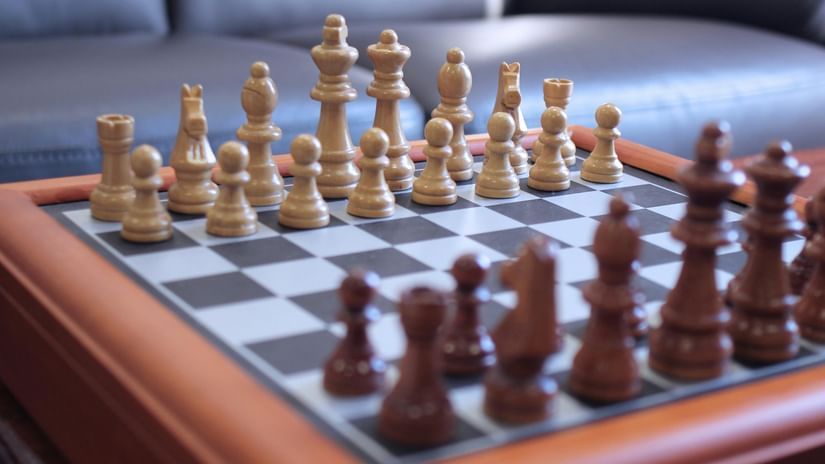 A chess set with all pieces arranged in the starting position on a wooden table, with a blurred sofa visible in the background.