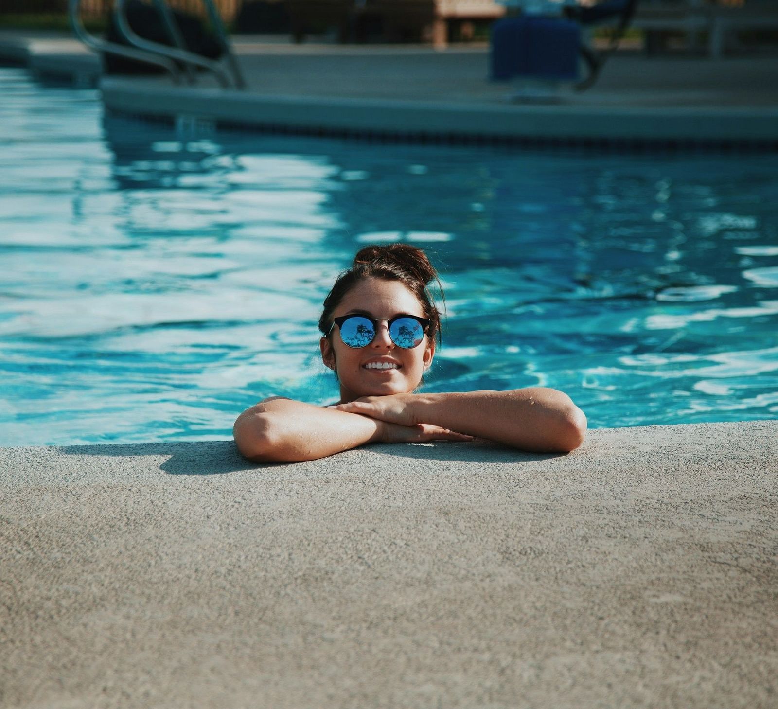 A girl in the swimming pool wearing sunglasses, with her hands resting on the floor, posing for a photo