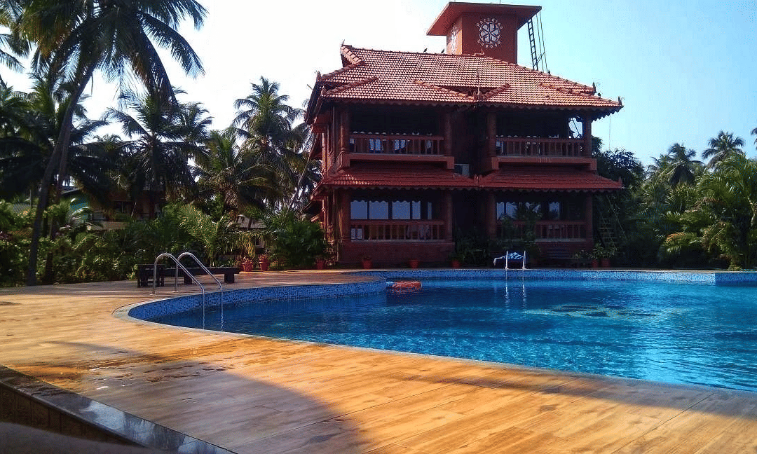 A large wooden building surrounded by palm trees with a clear blue swimming pool in the foreground at Paradise Lagoon Resort, Udupi.
