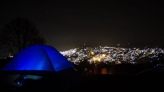A vibrant blue tent glows against the contrasting darkness of a clear, star-filled night sky, with a distant city’s lights shimmering below.