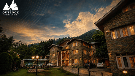 Exterior view of Hotel Outlook, Pahalgam, showing stone buildings next to a lawn, illuminated under a cloudy sky with hills in the background.
