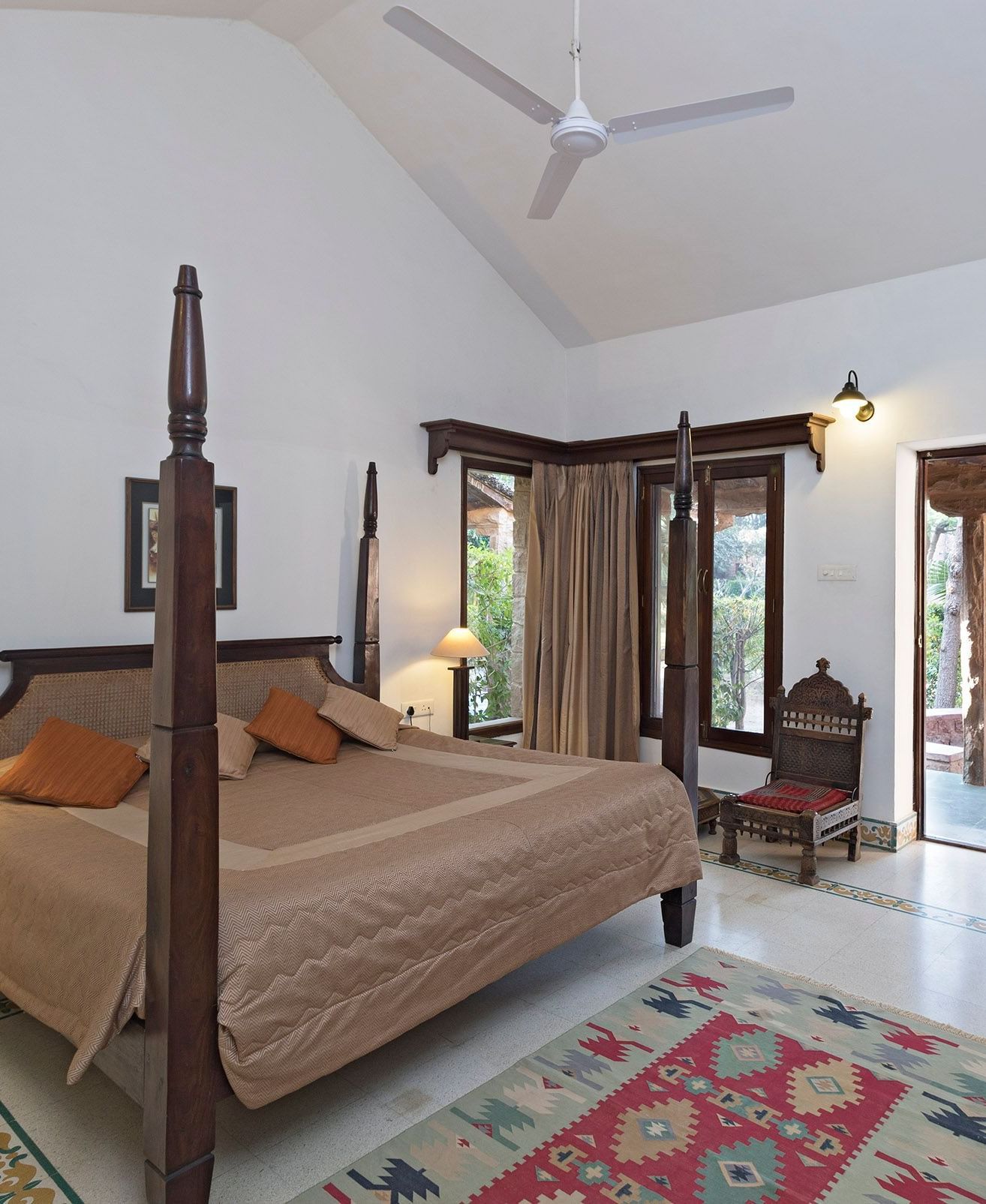 A bedroom with a four-poster double bed, wooden furniture, patterned rug, wardrobe, desk with kettle and cups, chair, ceiling fan, and doors opening outside - MANVÂR Shergarh The Desert Resort