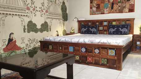 A Luxury Room at Jaypore360 Homestay with a wooden bed and nightstand with decorative tiles, a mural of traditional architecture and figures, and patterned pillows with camel motifs.