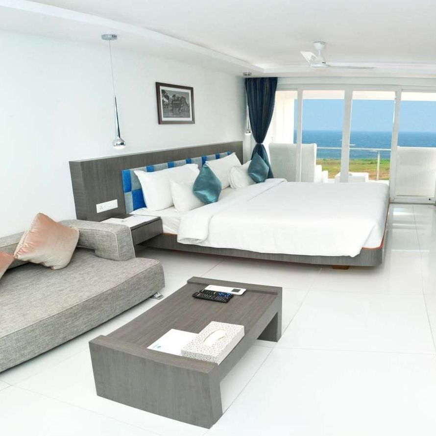A modern hotel room with a large bed, a grey sofa, and a coffee table, featuring floor-to-ceiling windows with blue curtains offering a view of the ocean | Grande Chalet Sea View | Grand Bay Resort and Spa