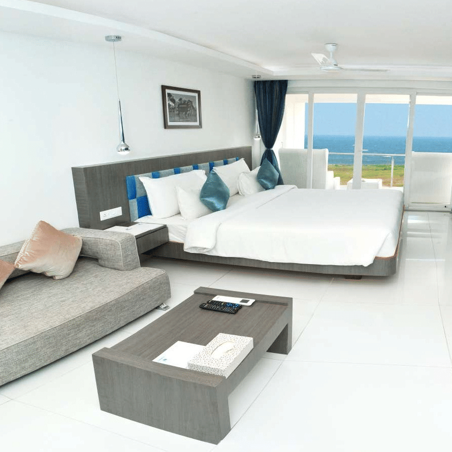 A modern hotel room with a large bed, a grey sofa, and a coffee table, featuring floor-to-ceiling windows with blue curtains offering a view of the ocean - Grande Bay Resort & Spa, Mamallapuram