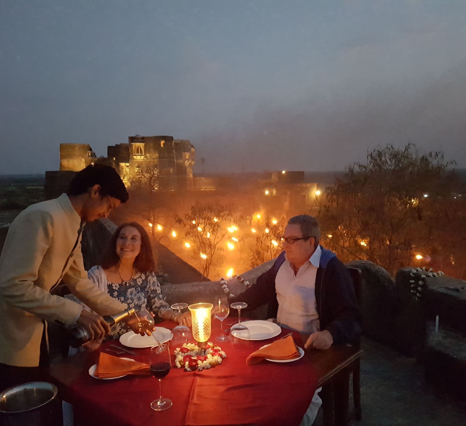 A waiter serves a meal to two people at a candlelit table on a rooftop at dusk, with lights visible in the distance- Shahpura Bagh.