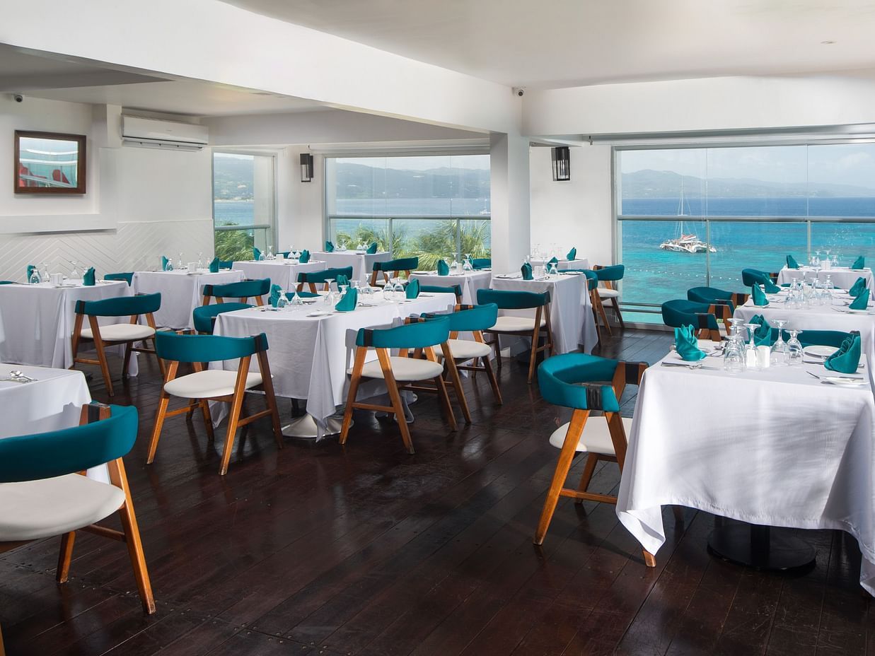 An interior view of a dining area at S Hotel Montego Bay with white tablecloths on tables, teal and wooden chairs, and large windows overlooking the ocean.