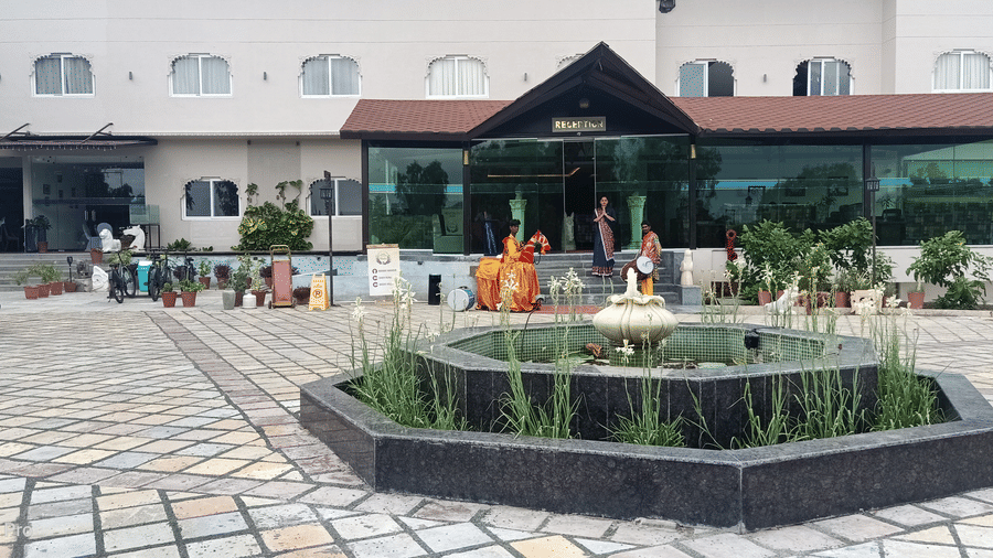 A facade view of a building with a covered entrance and a decorative fountain in a paved courtyard.