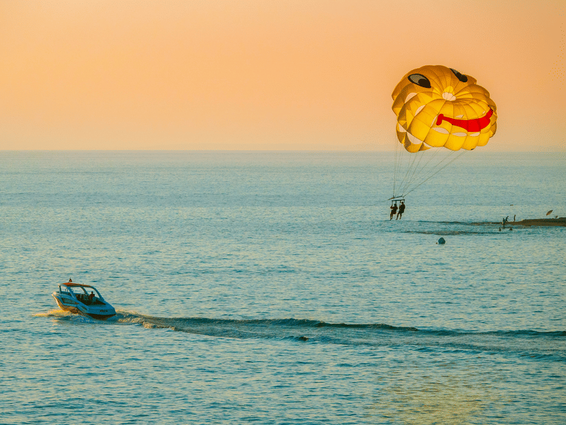 A wide view of 2 people parasailing above the ocean, hanging from a colourful parachute as a boat pulls them across the water.