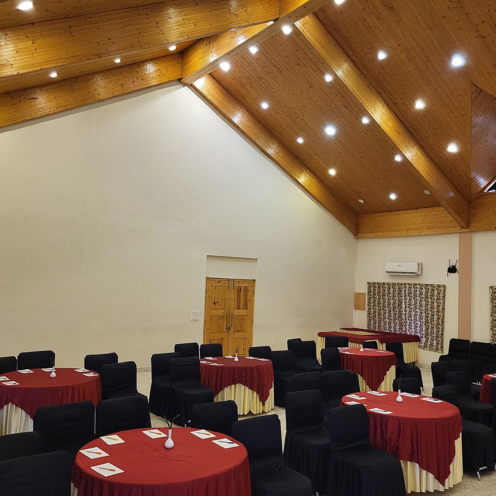 A spacious banquet hall with high wooden ceilings, elegant lighting, and round tables set for events at Dagshai Manor Kasauli - Inde Hotels.