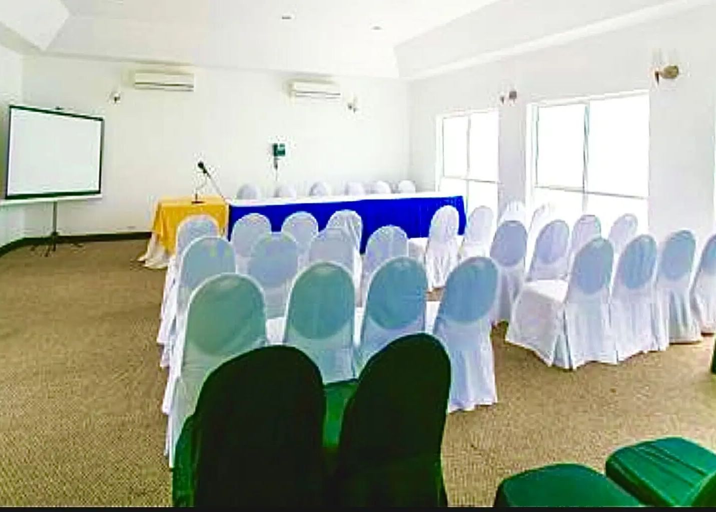 A meeting room is set up with rows of white chairs facing a table with a white tablecloth and a projector screen | Karnak | Altamont Court, Jamaica