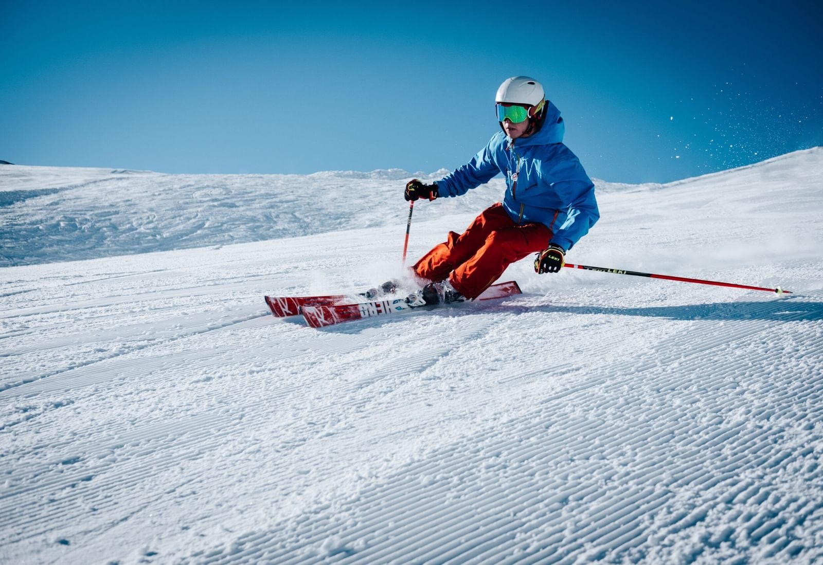 A landscape covered in snow featuring a person wearing a blue-colored jacket skiing through it with a clear blue sky in the background.