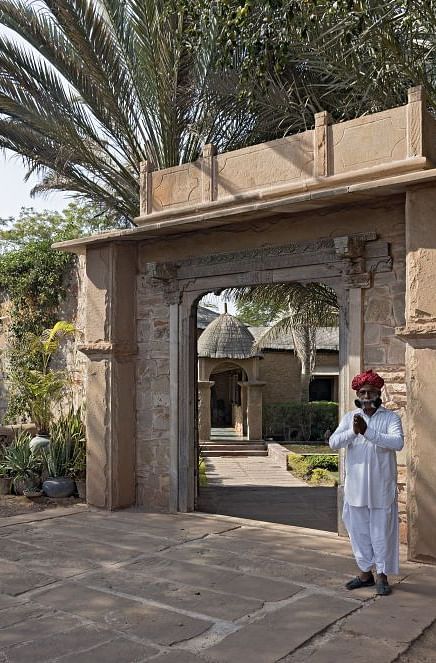 An image of a man in Rajasthani attire greeting with folded hands with the entrance to MANVAR Resort & Desert Camp in the background