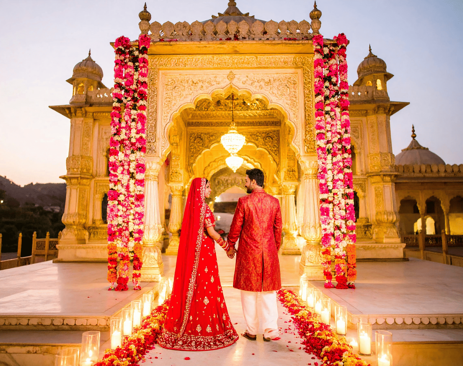 A bride and groom in red traditional attire pose intimately under an ornate stone archway decorated with red flowers and glowing lights.