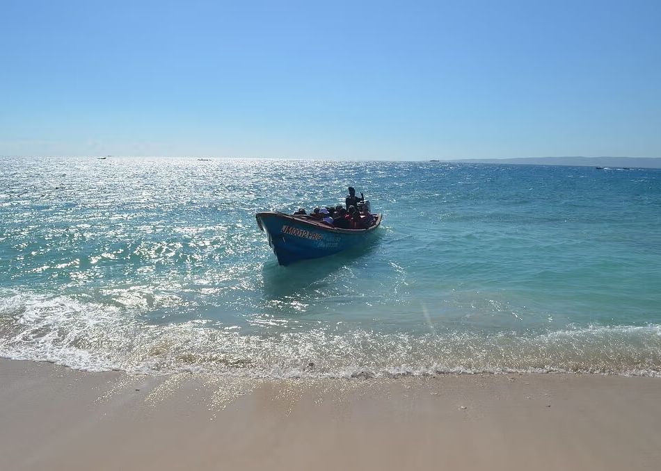 A small boat floats in calm, clear blue ocean water near a sandy beach under a bright sky.