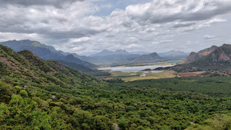 Panoramic view of the Palani Hills overlooking lush forests, rolling mountains, and a scenic reservoir under dramatic clouds.