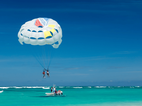 2 people parasailing, towed by a boat on ocean water beneath a bright blue sky.
