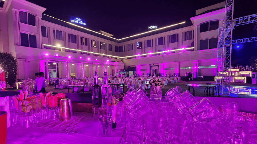 An event set up at the lawn at Ambrosia Sarovar Portico, Haridwar, featuring a bar counter with stacked glassware, illuminated with purple lighting and banquet tables.