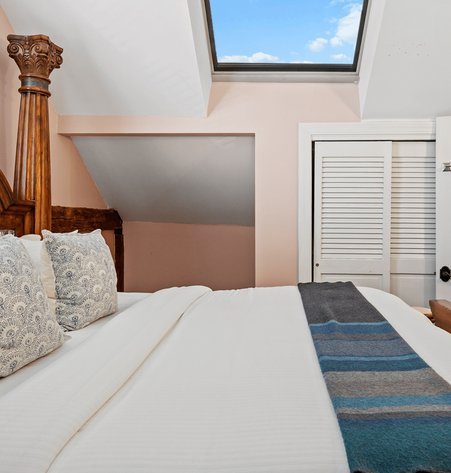 A neatly made bed with a wooden frame, pillows, and a throw blanket under a skylight in a room with peach walls.