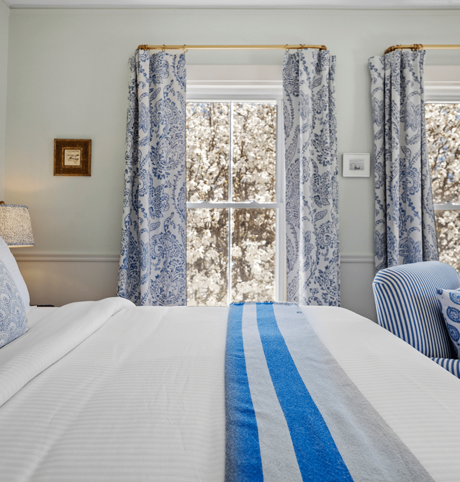 A neatly made bed with white bedding and blue accents in a room with floral curtains and a window view.