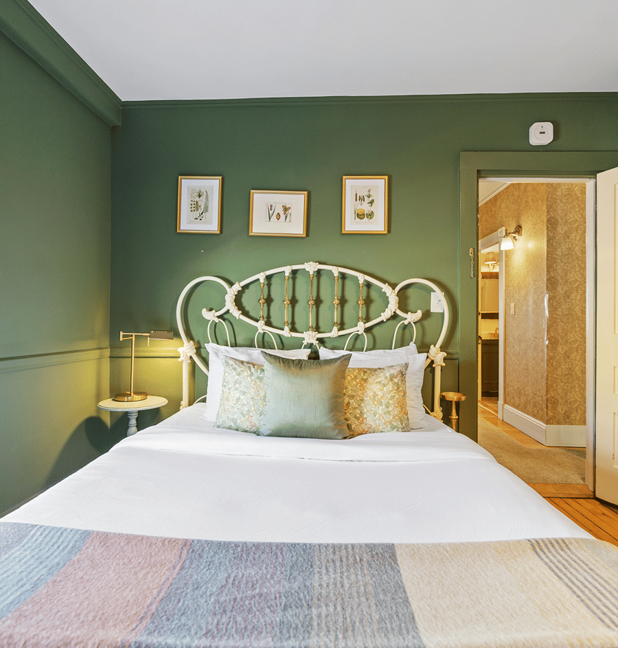 A neatly made bed with a white comforter and pillows, against a green wall with three framed pictures, with a doorway to another room in the background.