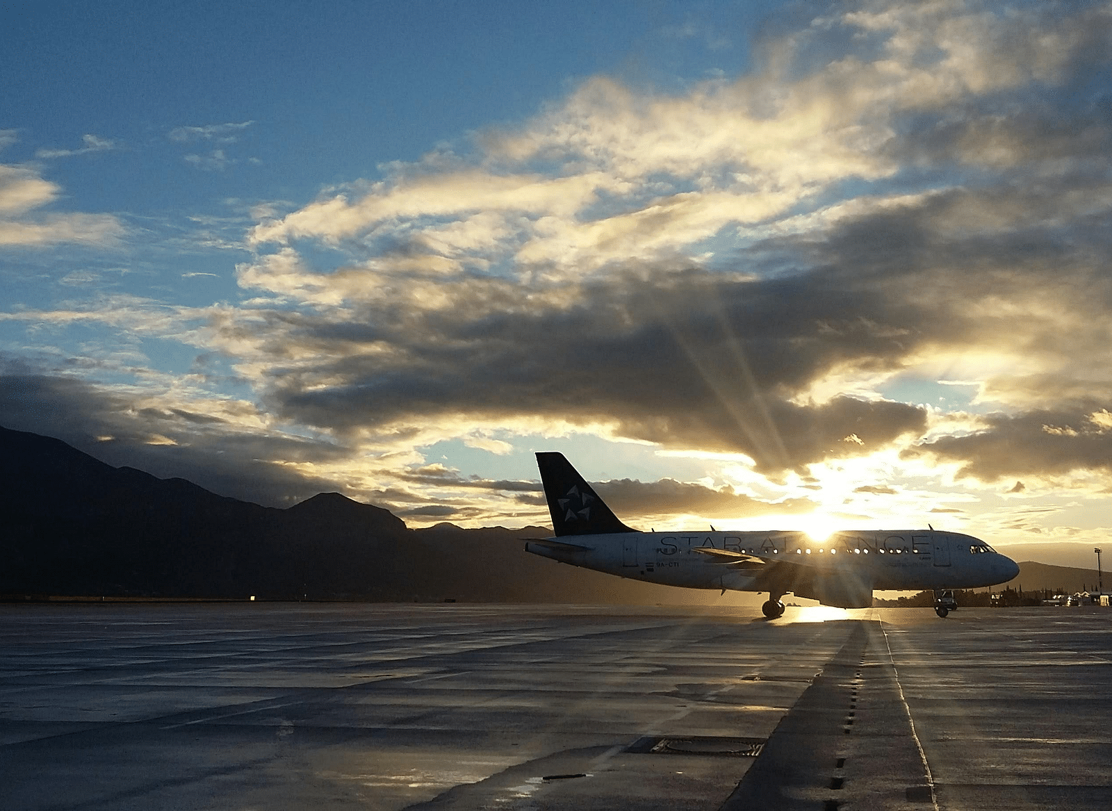 A commercial airplane is silhouetted against a dramatic sunset sky with dark, textured clouds, positioned on an airport tarmac that reflects the fading light, with a mountain visible in the distance.