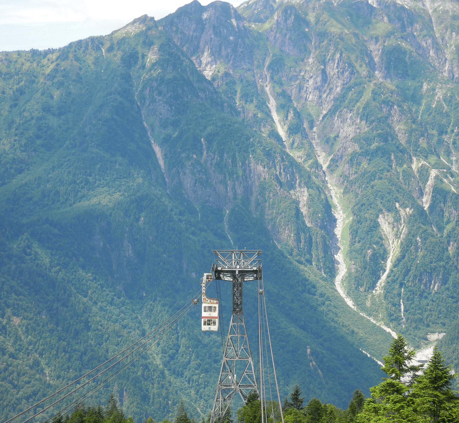 A cable car and support tower stand in front of a lush green mountain range.