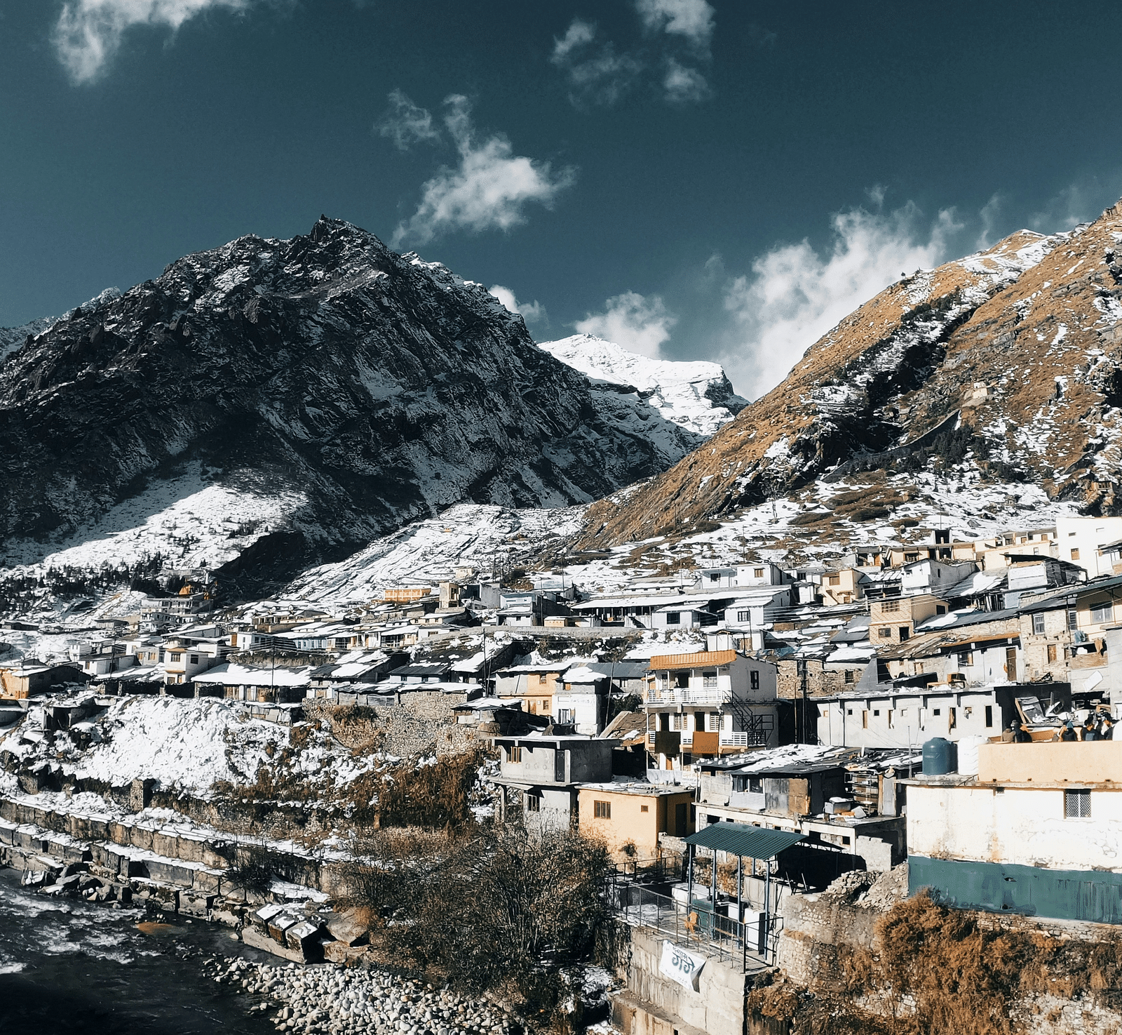 A snow-capped mountain looms over a village with tightly packed houses, beneath a dramatic sky.