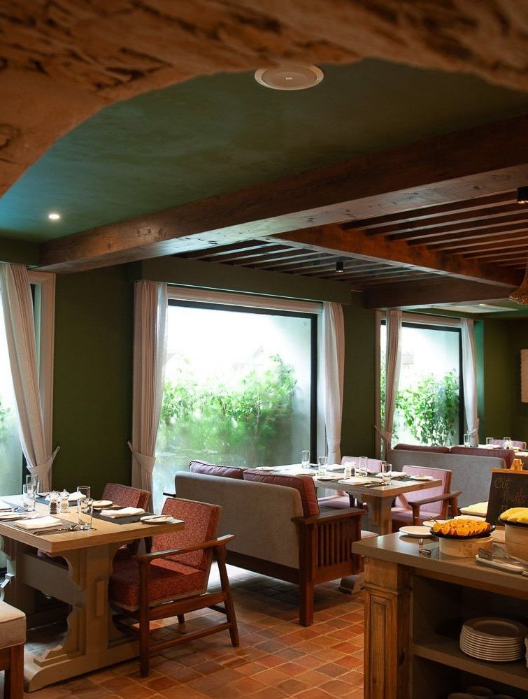 A wide-angle view of the dining area at Mila Restaurant at The Evren, Vagator, captured from a stone archway. The space features wooden tables, comfortable chairs and banquettes, and a buffet station with a variety of food.