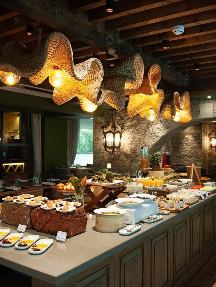 A lavish breakfast buffet spread at Mila Restaurant at The Evren, Vagator, featuring a variety of dishes on a large central island. The restaurant has a rustic yet modern ambiance with woven light fixtures, a stone archway, and a view of the outdoors.