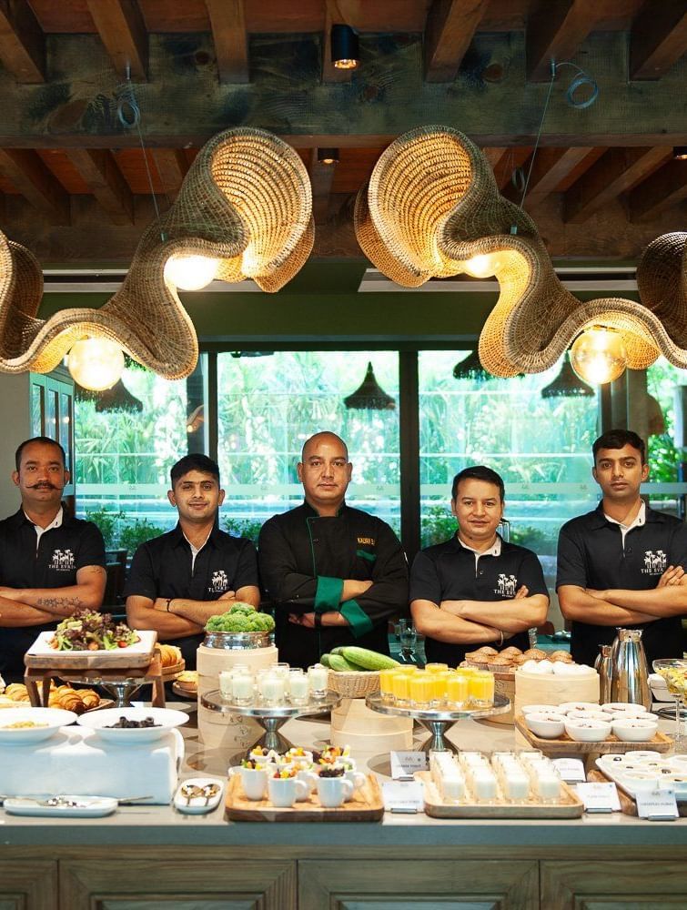  The culinary and service team of Mila Restaurant at The Evren, Vagator, standing proudly behind a beautifully presented buffet spread. The team is smiling and wearing their uniforms, with the head chef in the center.