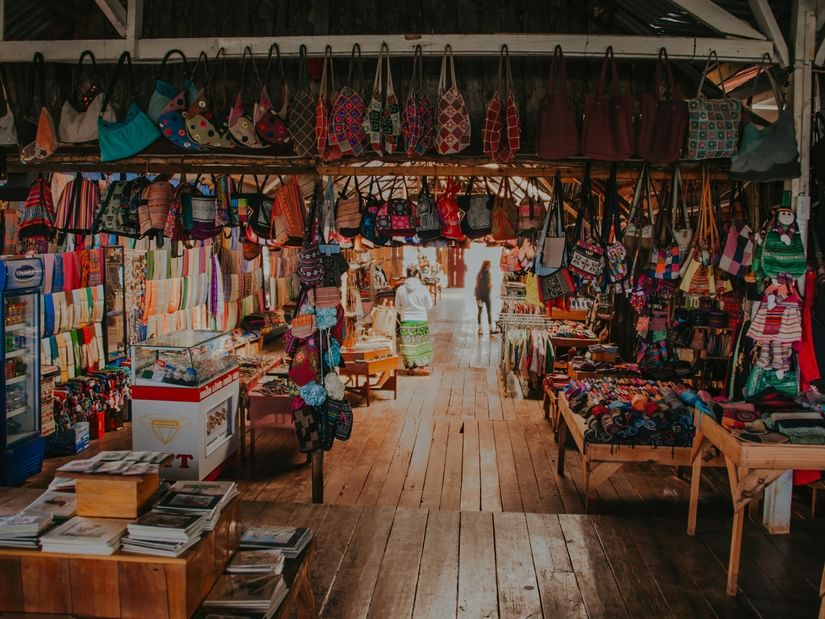 A vibrant indoor market stall is filled with colorful handbags and textiles hanging from the ceiling and displayed on wooden tables.