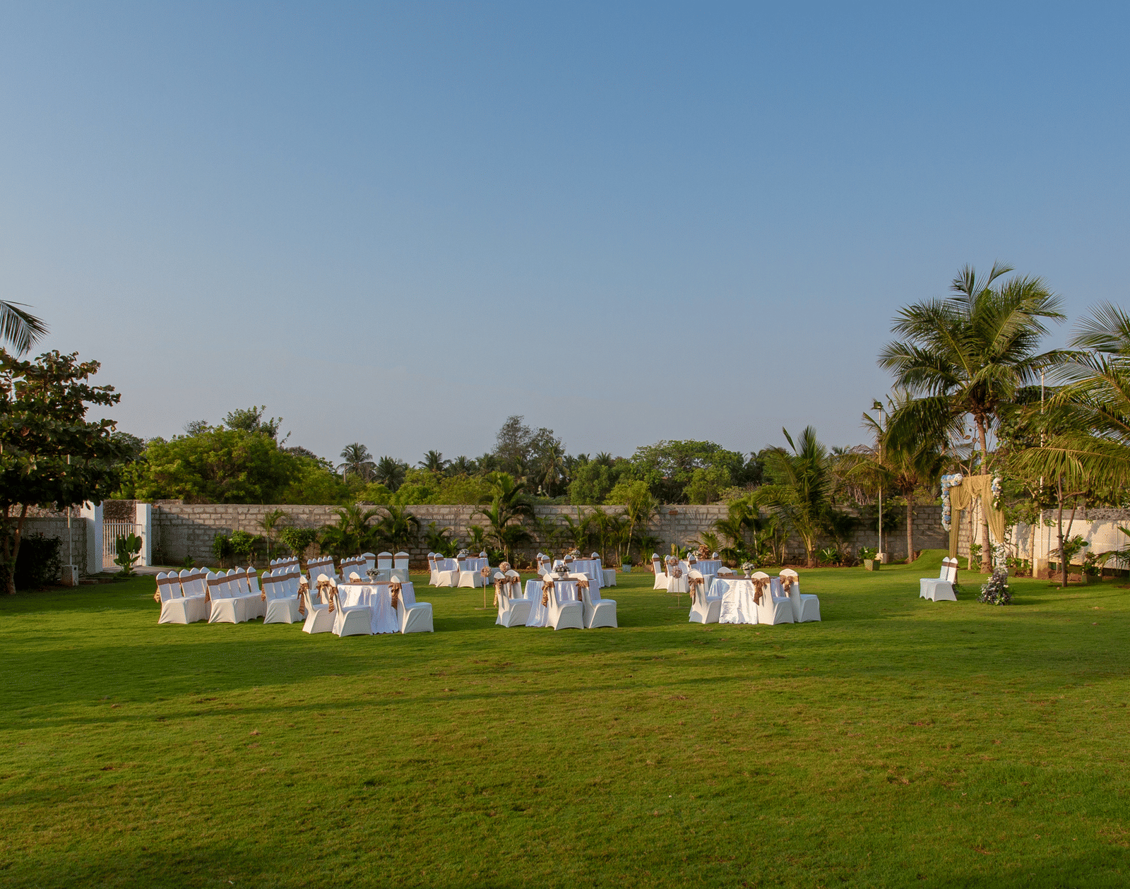 An outdoor event space with white chairs and tables arranged on a green lawn under a clear sky, with trees in the background - Grande Bay Resort & Spa, Mamallapuram