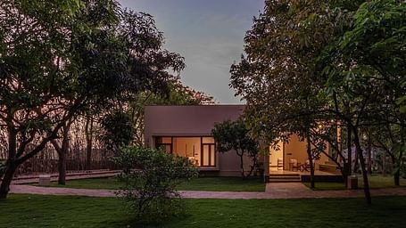 An exterior view of a building with lit windows, surrounded by trees and greenery at dusk - Shreyas Retreat
