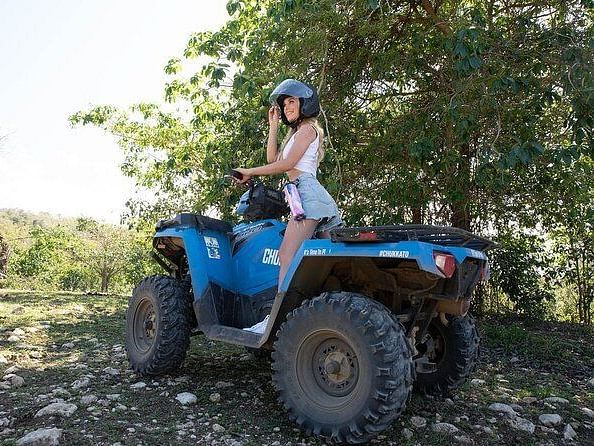 A person wearing a helmet sits on a quad bike in a natural, rocky setting with green foliage.