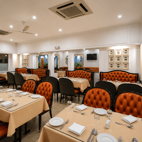 Tandoor restaurant with orange tufted banquettes, tables laid with cream cloths, framed butterfly displays and a marble floor at MAYFAIR Garden, Rourkela.