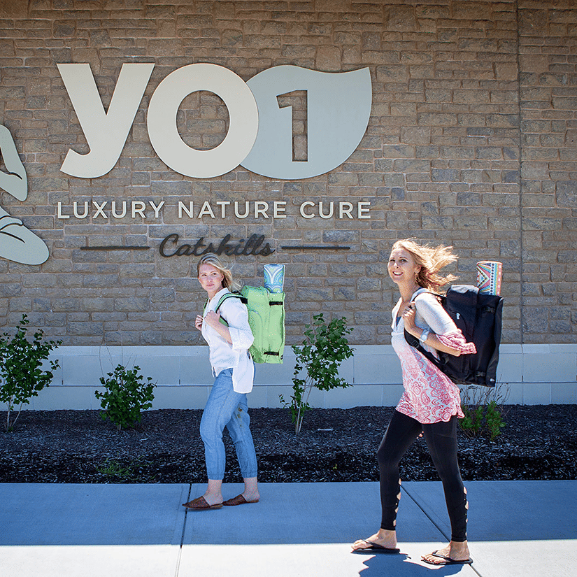 Health & Wellness Camp – Rebalance From Within at YO1 Longevity & Health Resorts - two guests carrying backpacks walking past the YO1 Luxury Nature Cure sign at the resort