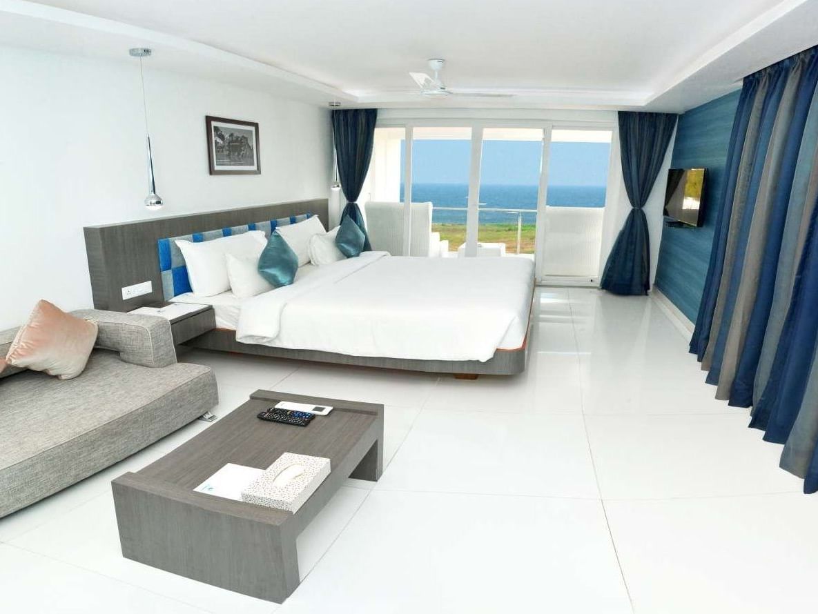 A modern hotel room with a large bed, a grey sofa, and a coffee table, featuring floor-to-ceiling windows with blue curtains offering a view of the ocean | Grande Chalet Sea View | Grand Bay Resort and Spa