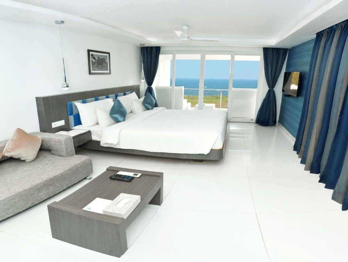 A modern hotel room with a large bed, a grey sofa, and a coffee table, featuring floor-to-ceiling windows with blue curtains offering a view of the ocean - Grande Bay Resort & Spa, Mamallapuram